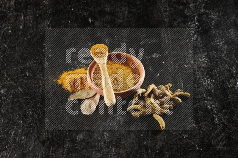 A wooden bowl and a wooden spoon full of turmeric powder with dried turmeric fingers on textured black flooring