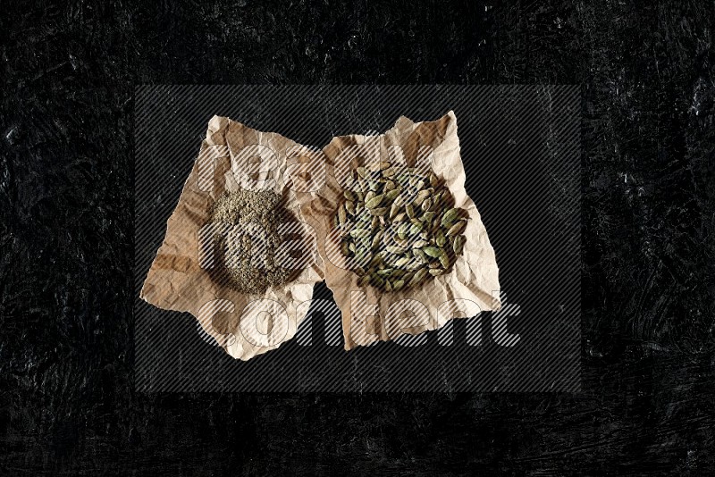 Cardamom seeds and cardamom powder in 2 crumpled pieces of paper on textured black flooring