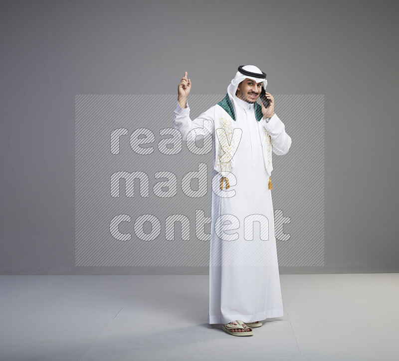 A saudi man standing wearing thob and white shomag with flag scarf on his neck talking on phone on gray background