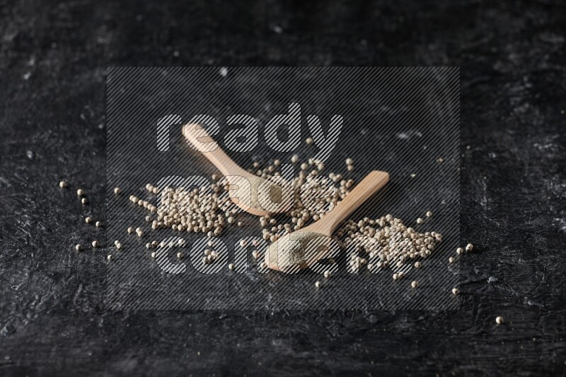 2 wooden spoons full of white pepper powder with white pepper beads on textured black flooring