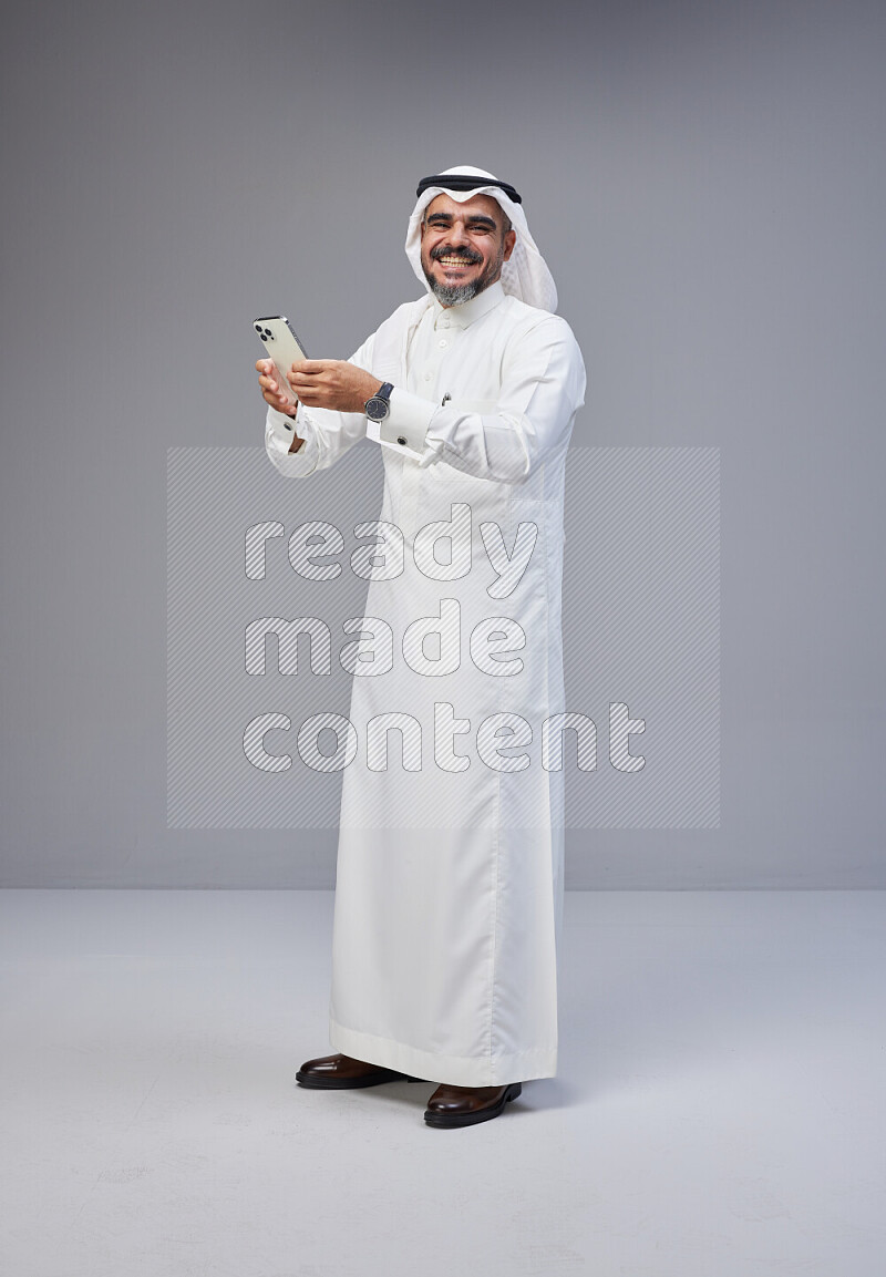 Saudi man Wearing Thob and white Shomag standing texting on phone on Gray background