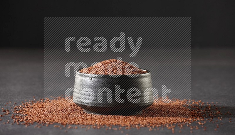 A black pottery bowl full of garden cress seeds surrounded by seeds on a black flooring