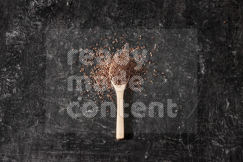 A wooden spoon full of flaxseeds and surrounded by seeds on a textured black flooring
