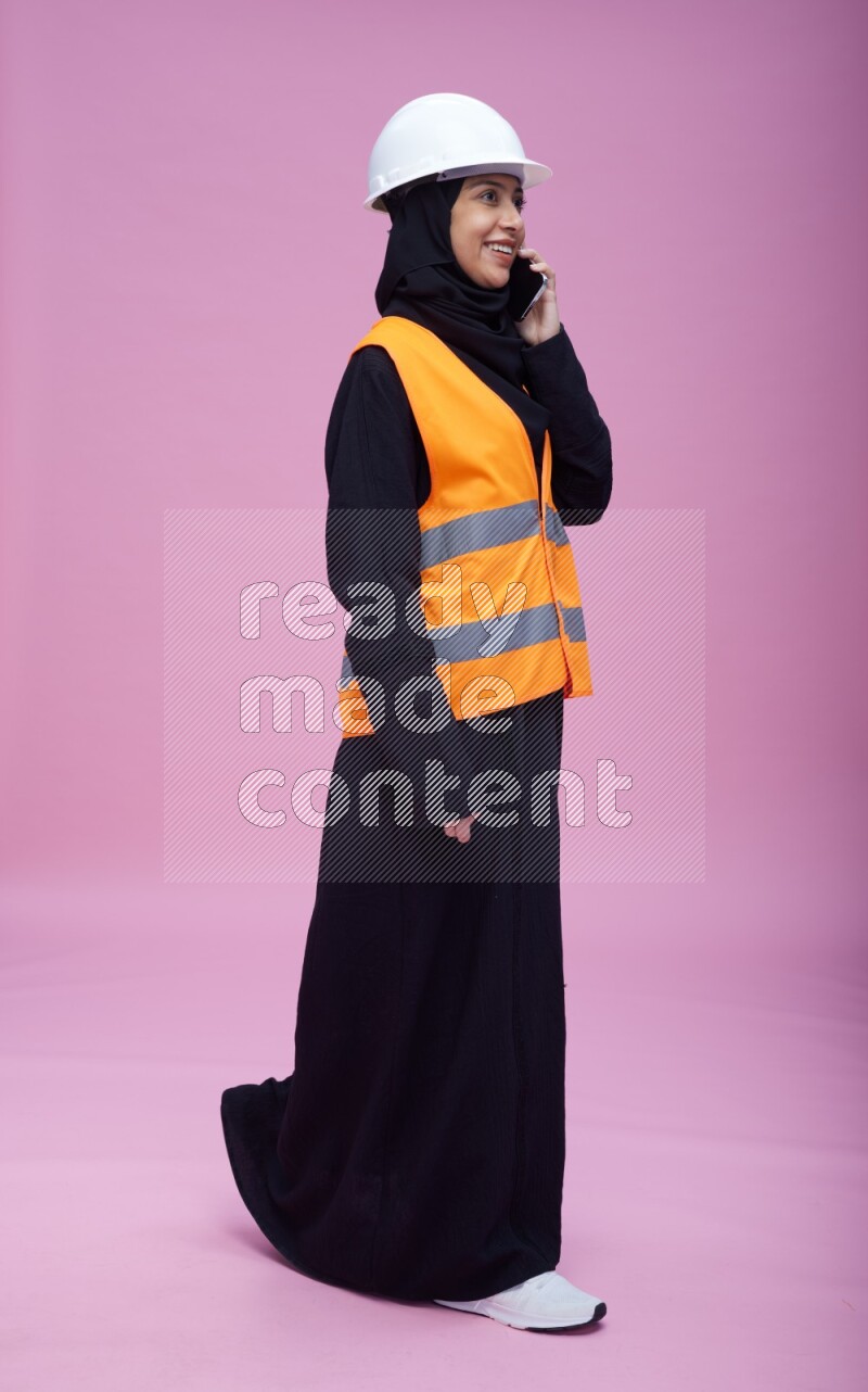 Saudi woman wearing Abaya with engineer vest and helmet standing talking on phone on pink background