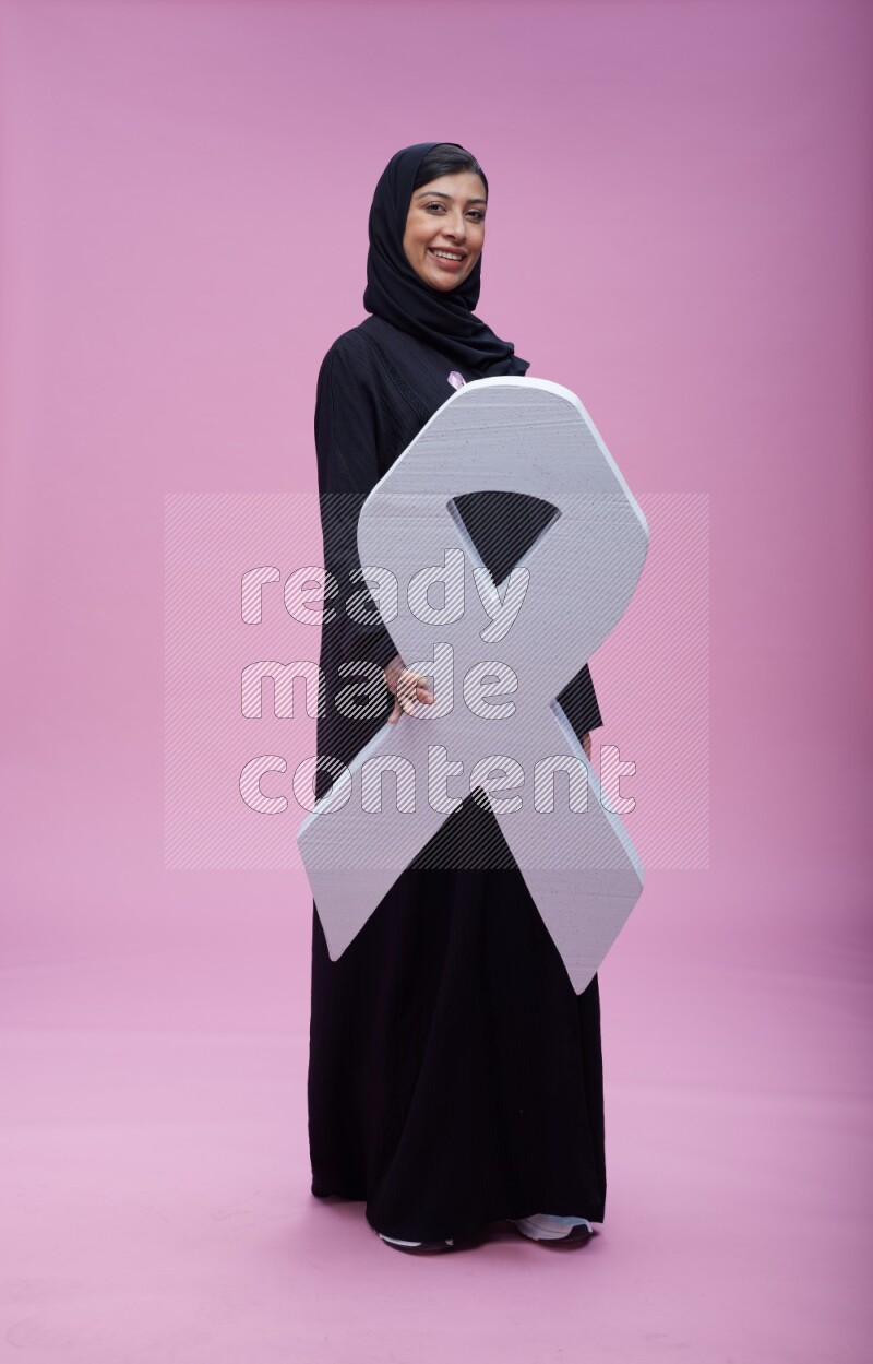Saudi woman wearing pink ribbon on abaya standing holding awareness ribbon on plain pink background