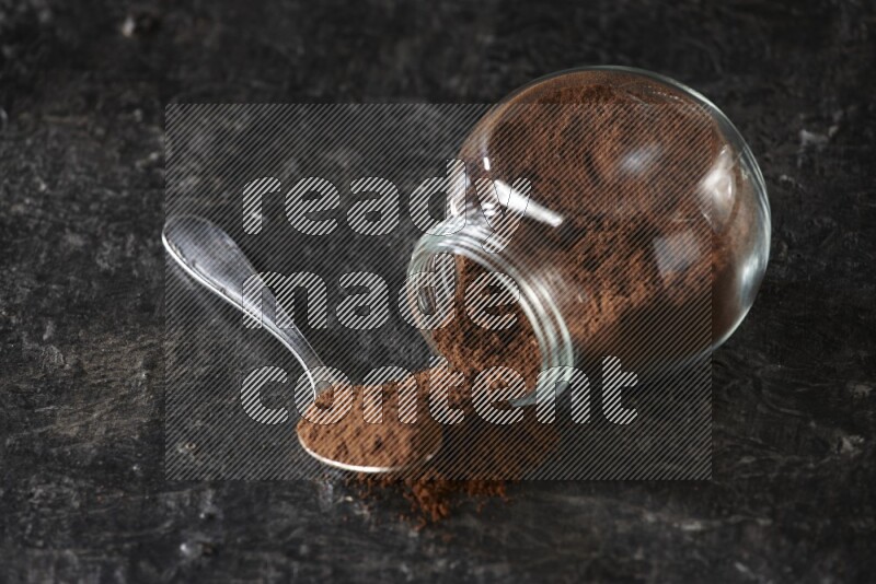 A flipped glass spice jar and a metal spoon full of cloves powder on textured black flooring