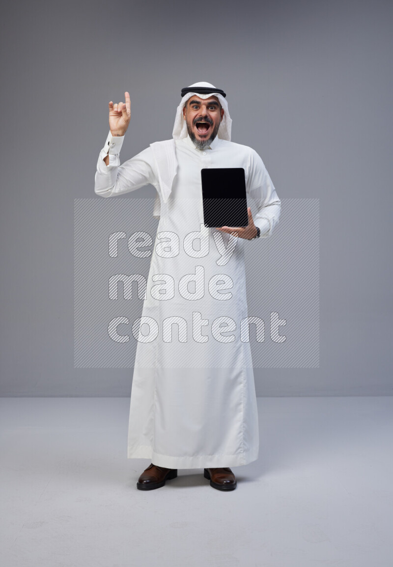Saudi man Wearing Thob and white Shomag standing showing tablet to camera on Gray background