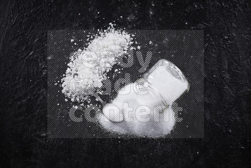 A glass jar full of table salt with some sea salt crystals beside it on a black background