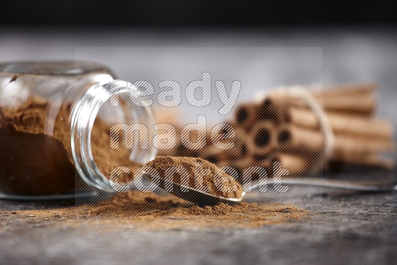 Herbal glass jar full cinnamon powder flipped and a metal spoon full of powder, cinnamon sticks stacked and bounded in the back on textured black background in different angles