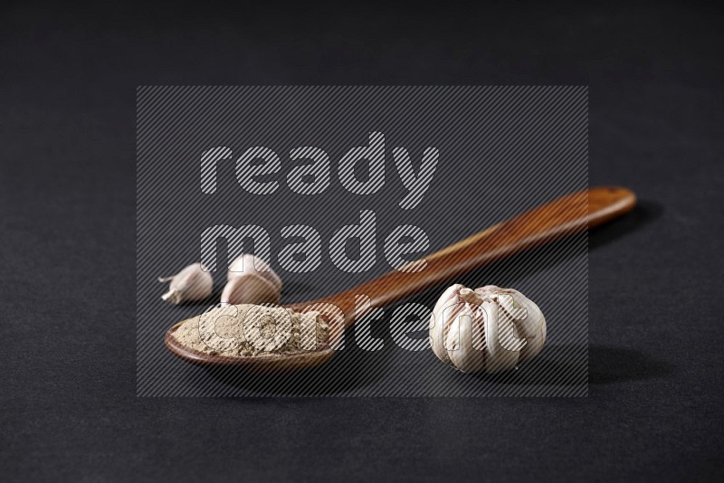 A wooden ladle full of garlic powder with garlic bulb and cloves beside it on a black flooring in different angles