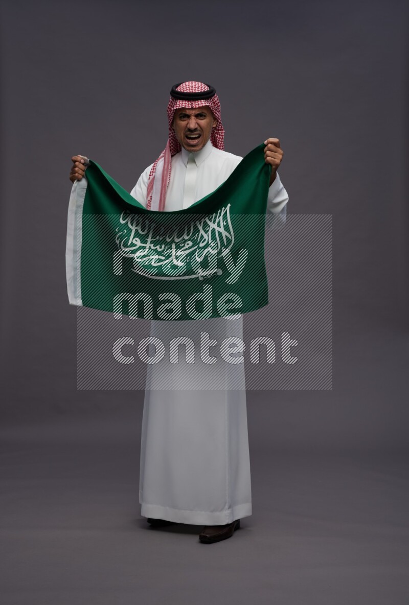 Saudi man wearing thob and shomag standing holding Saudi flag on gray background