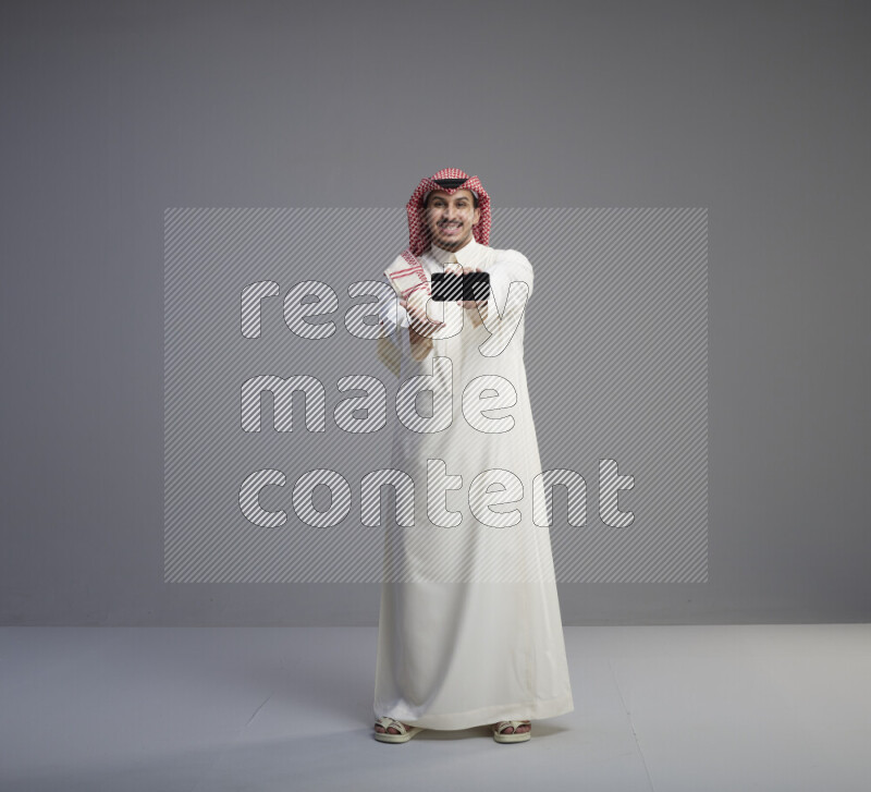 A Saudi man standing wearing thob and red shomag showing phone to camera on gray background