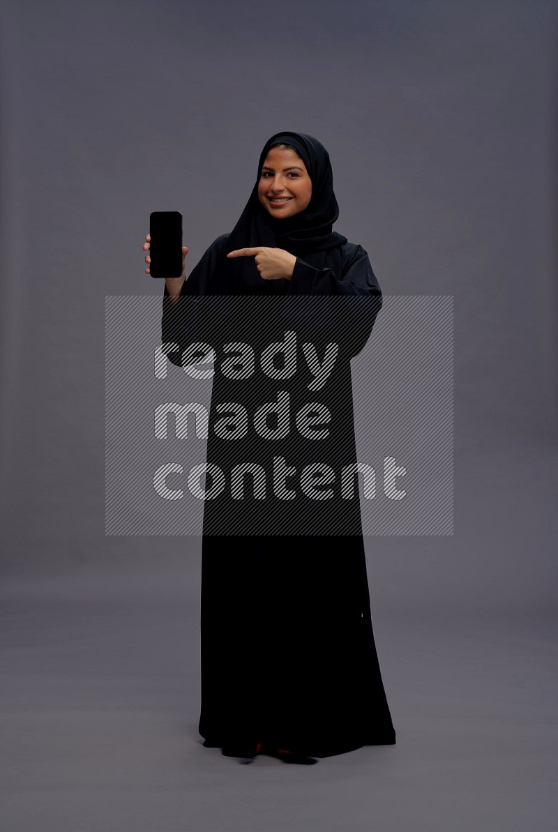 Saudi woman wearing Abaya standing showing phone to camera on gray background