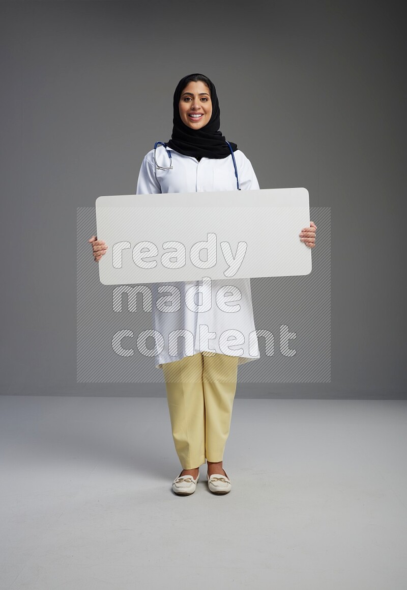 Saudi woman wearing lab coat with stethoscope standing holding board on Gray background