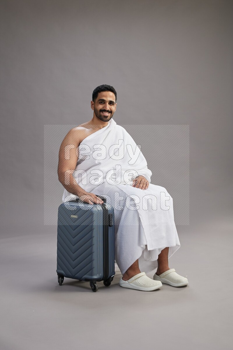 A man wearing Ehram sitting on chair holding traveling bag on gray background