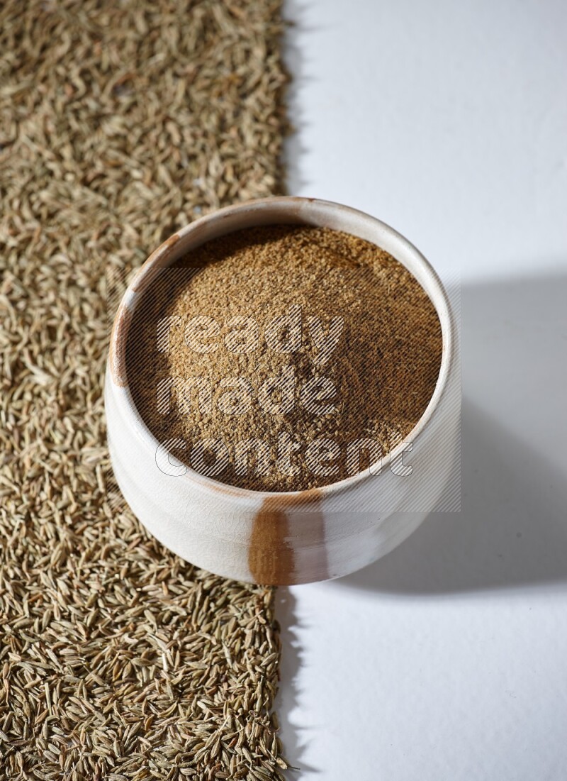 A beige bowl full of cumin powder with cumin seeds beneath it the bowl on a white flooring