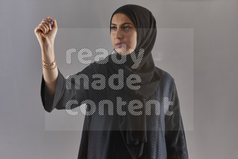 Woman in black abaya and hijab holding a marker pen to write on a transparent board with different reactions and expressions on grey background