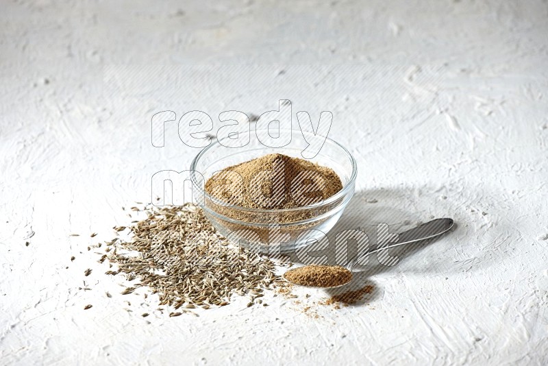 A glass bowl and metal spoon full of cumin powder and cumin seeds underneath it on textured white flooring