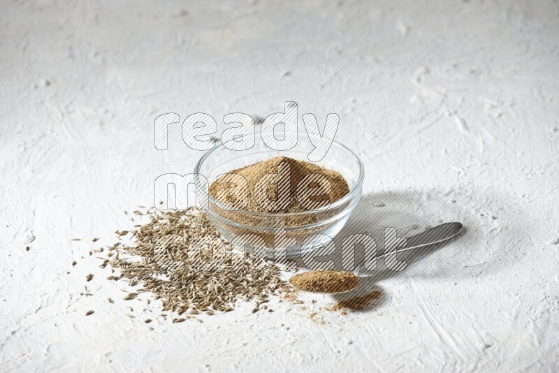 A glass bowl and metal spoon full of cumin powder and cumin seeds underneath it on textured white flooring
