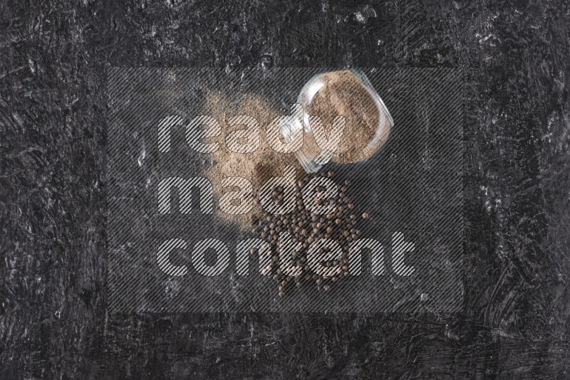 A flipped glass spice jar full of black pepper powder with pepper beads on textured black flooring