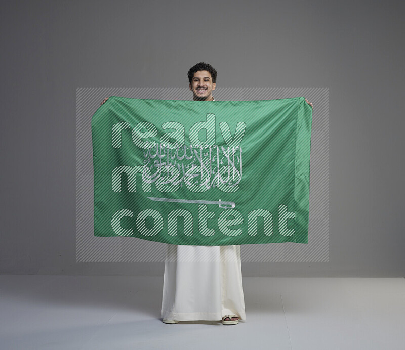 A Saudi man standing wearing thob holding big Saudi flag on gray background