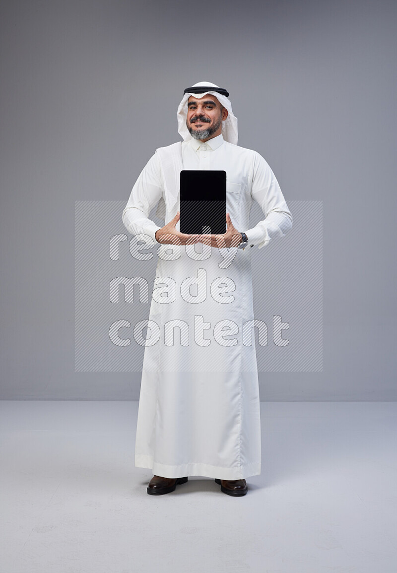 Saudi man Wearing Thob and white Shomag standing showing tablet to camera on Gray background