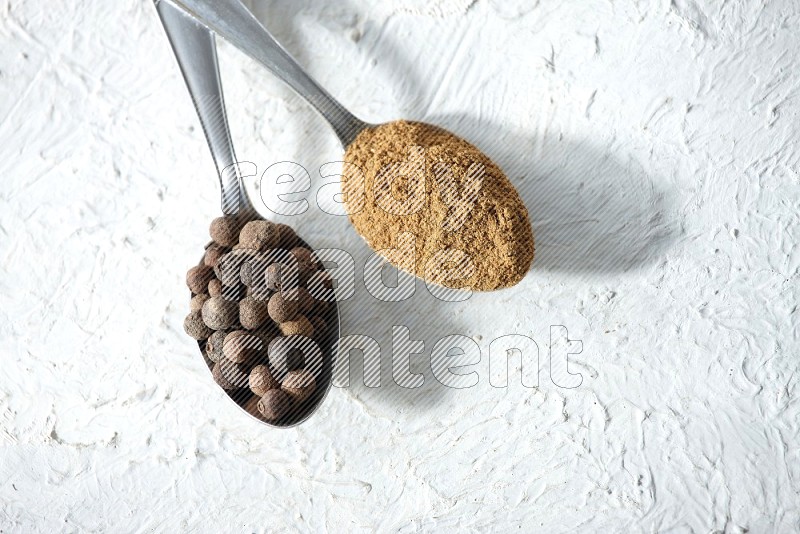 2 metal spoons full of allspice powder and whole balls on a textured white flooring