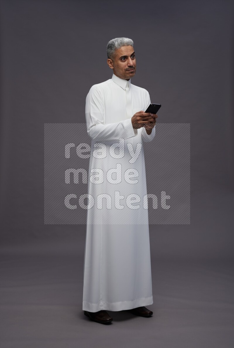 Saudi man wearing thob standing texting on phone on gray background