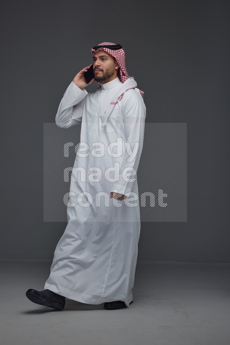 A Saudi man wearing Thobe and Shmagh talking in the phone eye level on a gray background