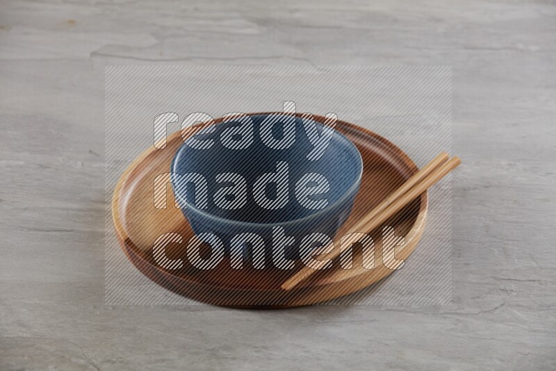 Blue pottery round bowl on top of brown wood round plate and wood chopsticks, on grey textured countertop