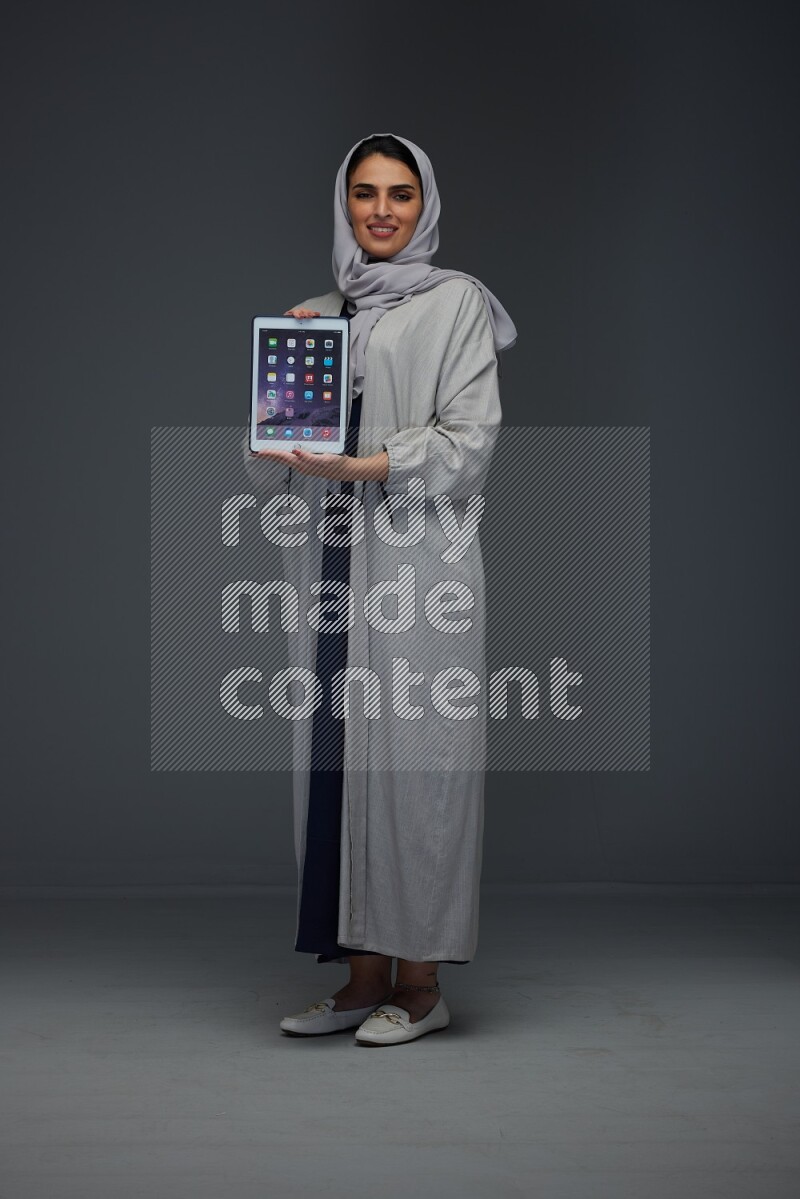 A Saudi woman wearing a light gray Abaya and head scarf standing and showing the phone's screen eye level on a grey background