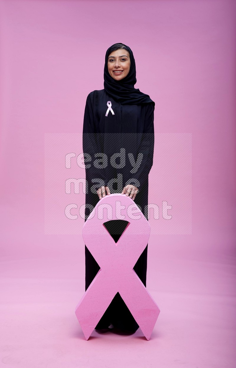 Saudi woman wearing pink ribbon on abaya standing holding awareness ribbon on plain pink background