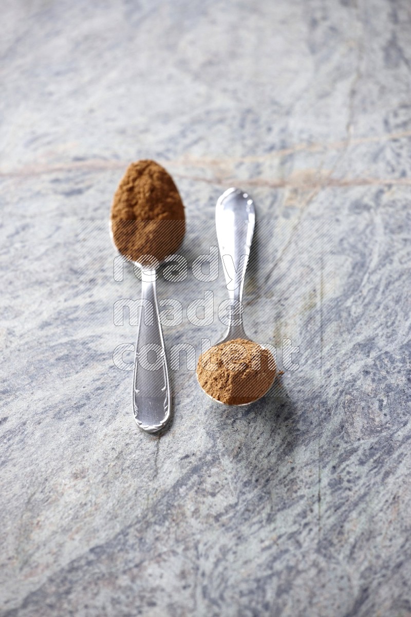 Two metal spoons full of cinnamon powder on grey marble background
