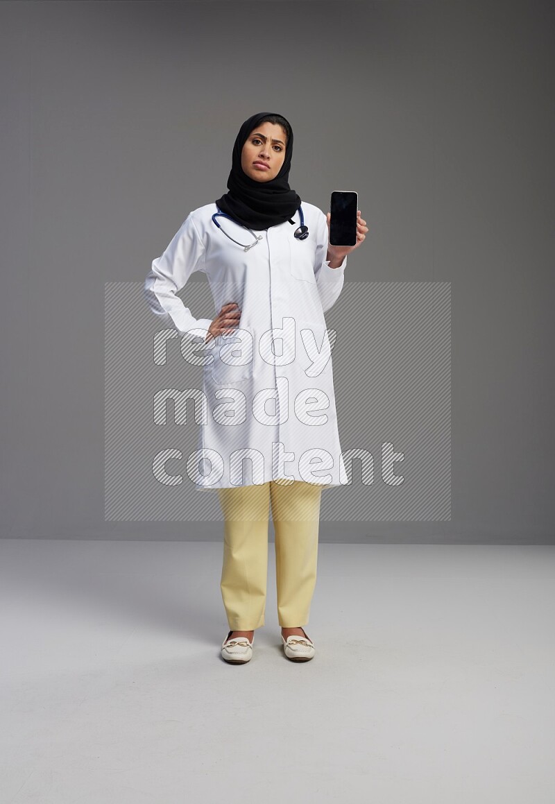 Saudi woman wearing lab coat with stethoscope standing showing phone to camera with sign in the back on Gray background
