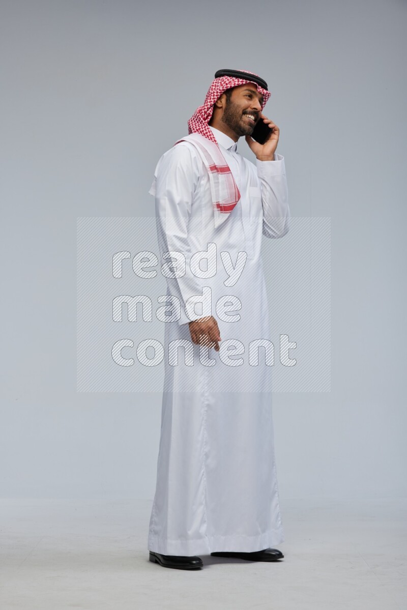 Saudi man Wearing Thob and shomag standing talking on phone on Gray background