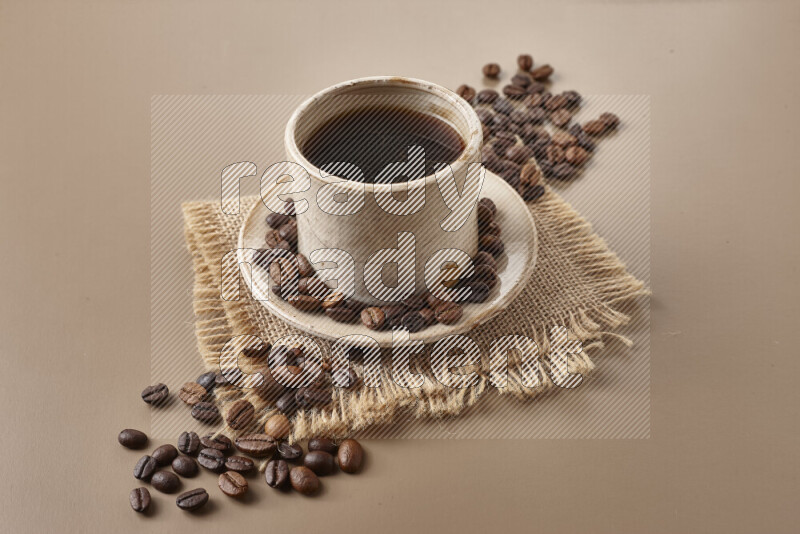 A beige pottery cup of coffee surrounded by roasted coffee beans on beige background
