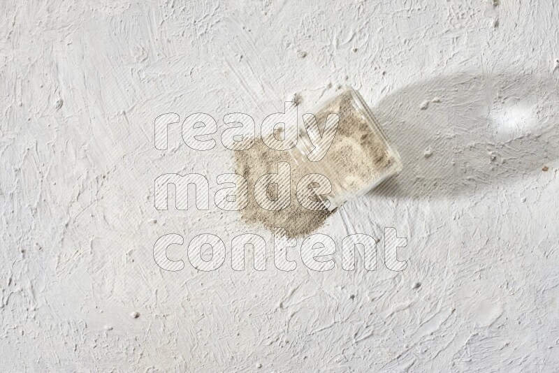 A flipped glass jar full of white pepper powder with spilled powder on textured white flooring