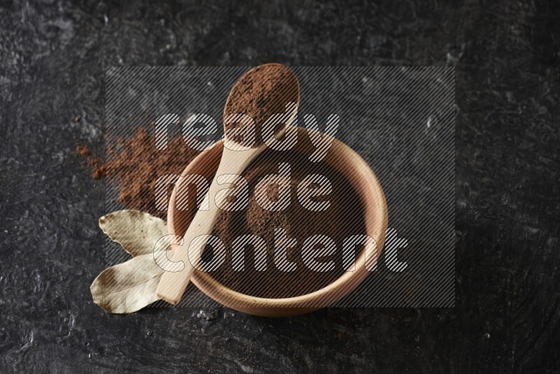 A wooden bowl and a wooden spoon full of cloves powder with laurel leaves on a textured black flooring