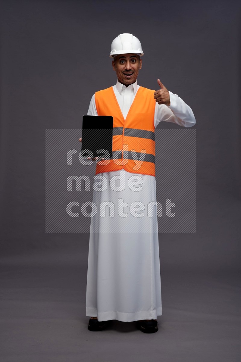 Saudi man wearing thob with engineer vest standing showing tablet to camera on gray background