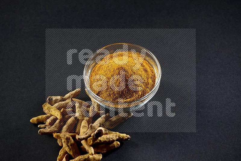A glass bowl full of turmeric powder and dried whole fingers beside it on black flooring