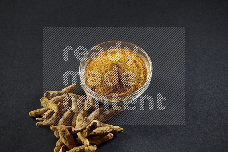 A glass bowl full of turmeric powder and dried whole fingers beside it on black flooring