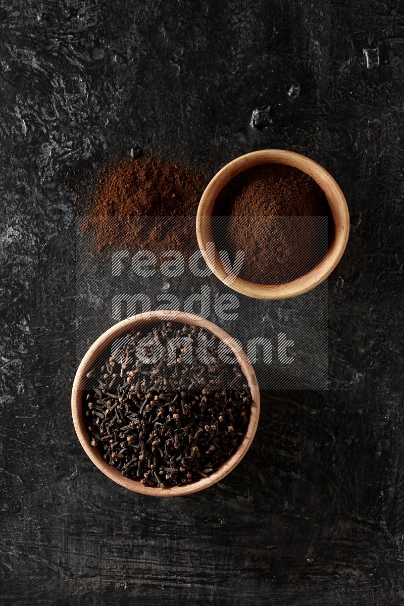 2 wooden bowls full of cloves powder and whole cloves on a textured black flooring