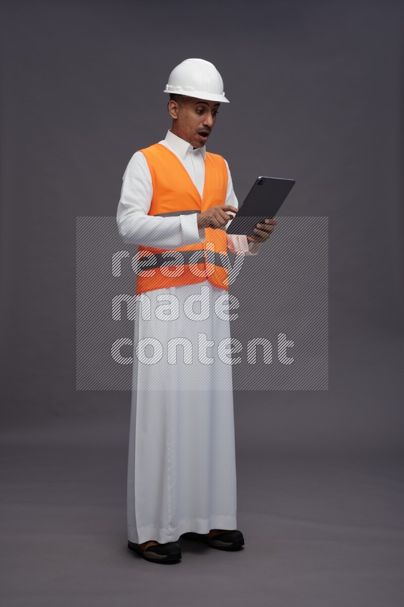 Saudi man wearing thob with engineer vest standing working on tablet on gray background