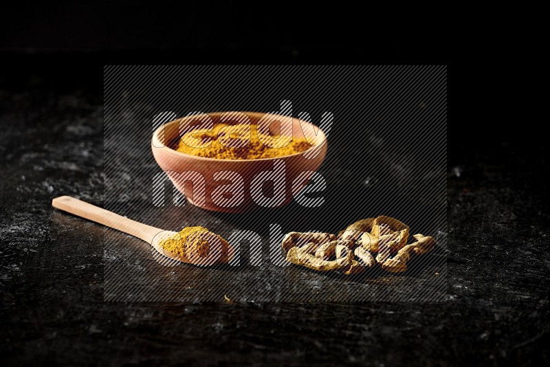 A wooden bowl and a wooden spoon full of turmeric powder with dried turmeric fingers on textured black flooring