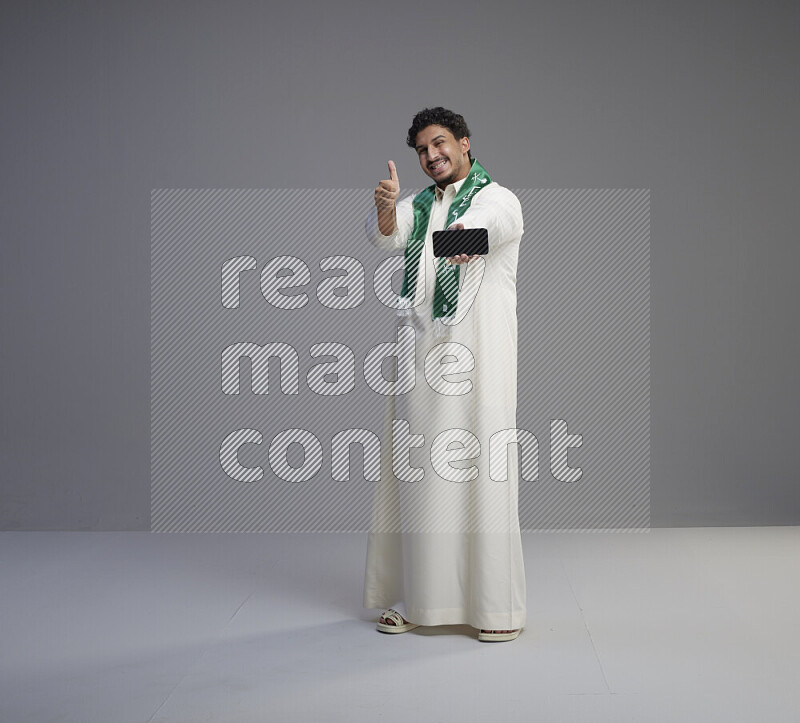 A Saudi man standing wearing thob and Saudi flag scarf showing phone to camera on gray background