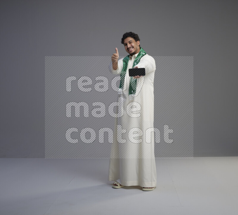 A Saudi man standing wearing thob and Saudi flag scarf showing phone to camera on gray background