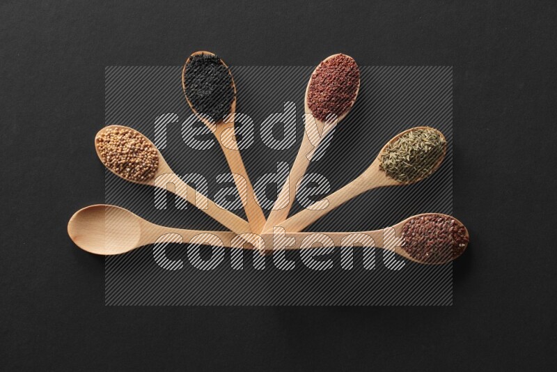wooden spoons filled with white peppers, mustard seeds, black seeds, garden cress, cumin and flax on black flooring
