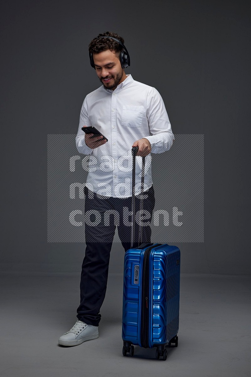 A man wearing smart casual using his phone while wearing headphone and holding luggage eye level on a gray background