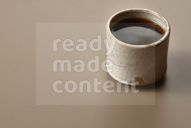 A beige pottery cup of coffee surrounded by roasted coffee beans on beige background