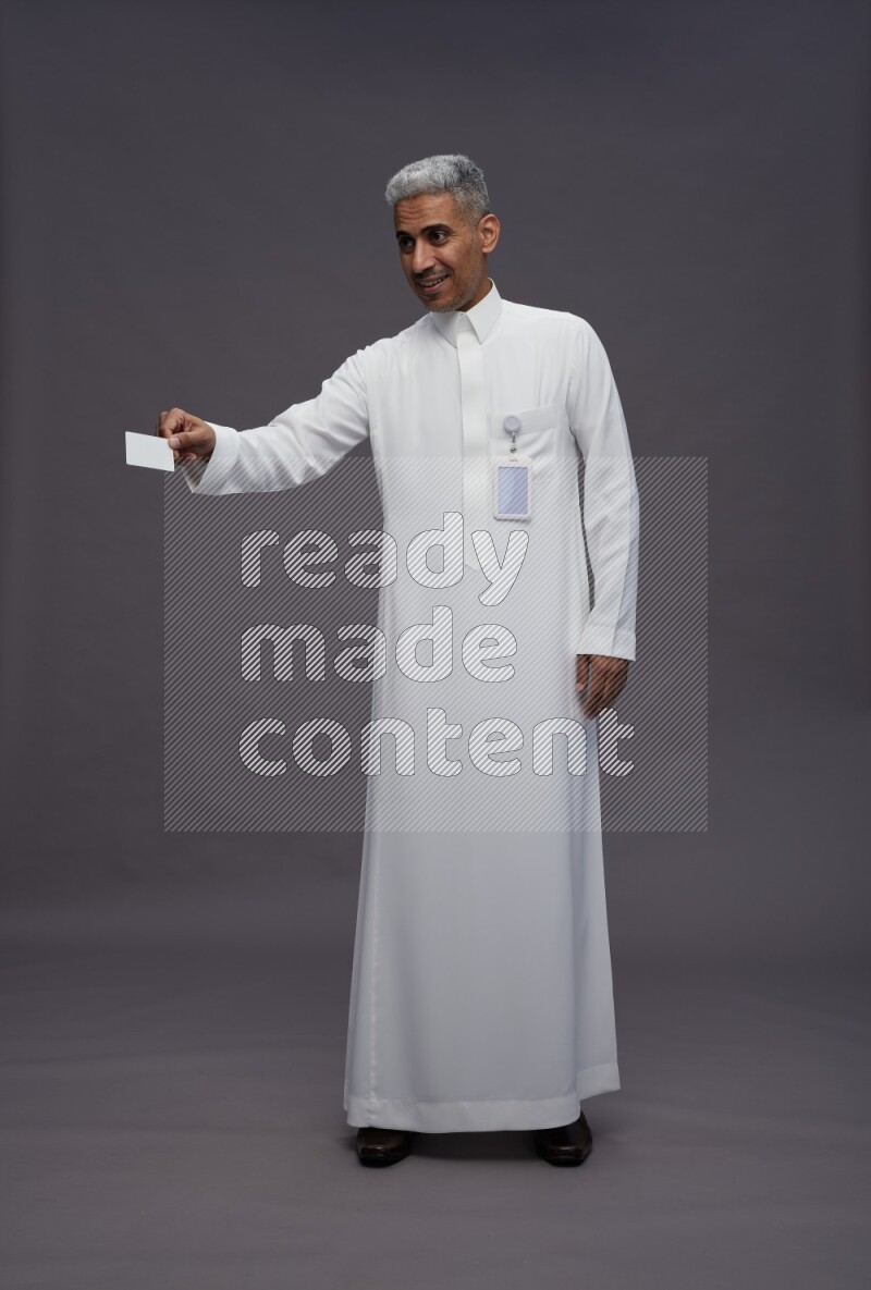 Saudi man wearing thob with pocket employee badge standing holding ATM card on gray background